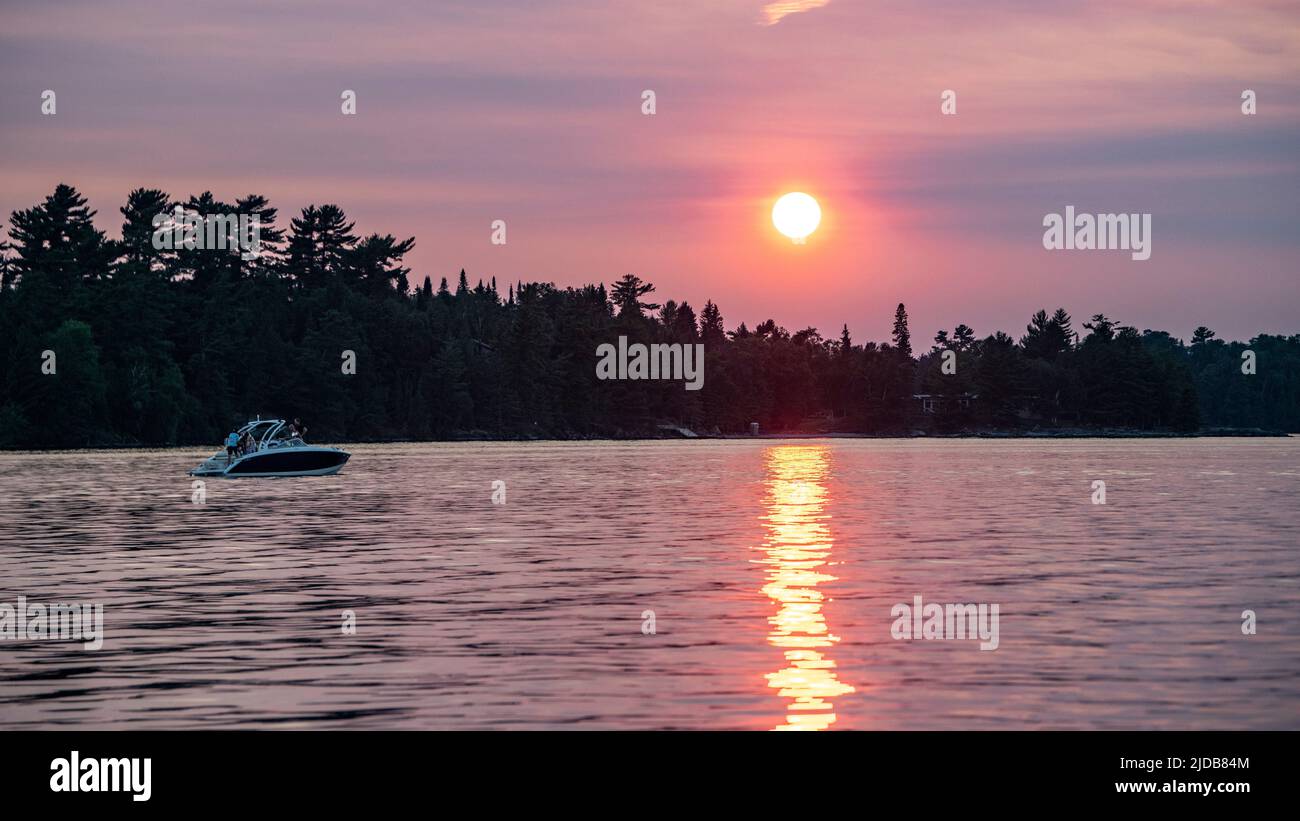 In barca su un lago al tramonto con un sole rosa vibrante che risplenderà su paesaggi e acqua silhouette, Lake of the Woods, Ontario; Kenora, Ontario, Canada Foto Stock