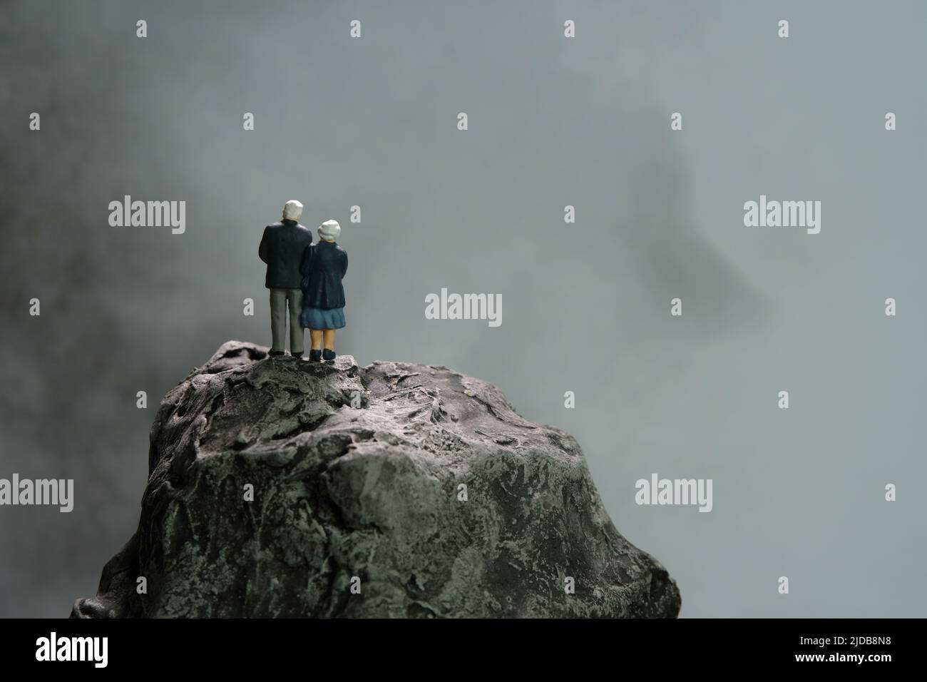 Persone in miniatura giocattoli fotografia concettuale. Coppia anziana in piedi sopra la roccia di montagna a tempo di pioggia nuvoloso scuro, pensando al futuro, vivendo, Foto Stock