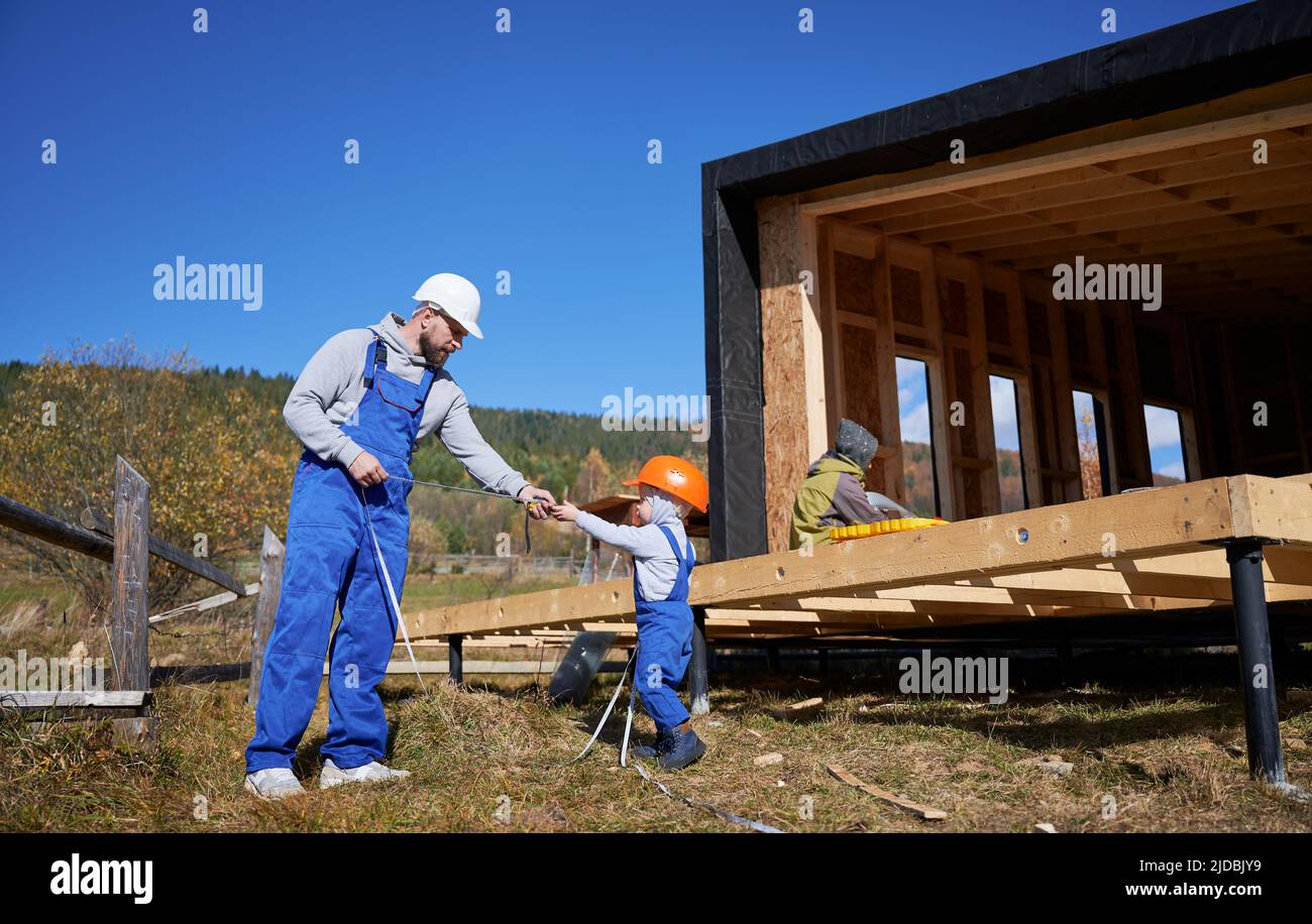 Padre con bambino figlio costruzione di legno telaio casa su palo fondazione. Ragazzo che aiuta il suo papà, giocando con metro a nastro sul cantiere, indossando casco e tute blu. Concetto di carpenteria. Foto Stock