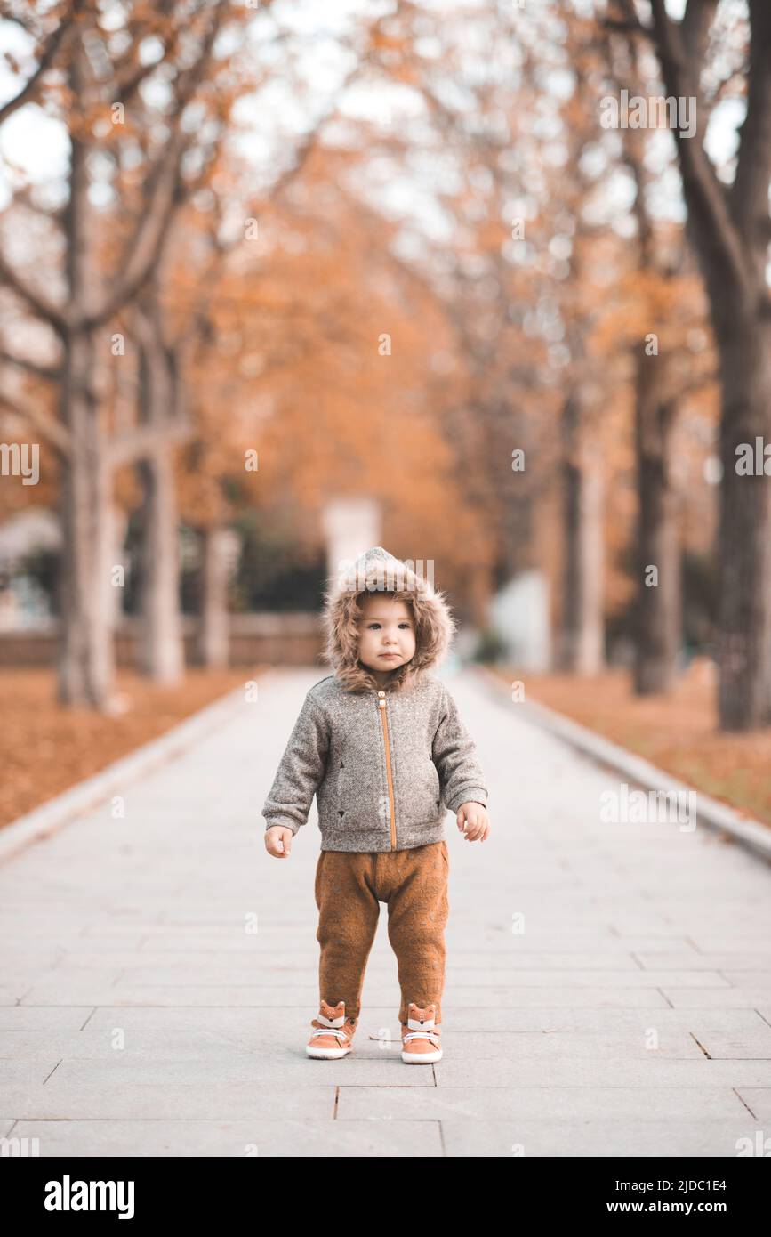 Cute baby girl 1-2 anni indossare giacca con cappuccio e pantaloni a piedi nel parco autunno sopra le foglie cadute e la natura all'aperto. Stagione autunnale. Infanzia. Poco Foto Stock