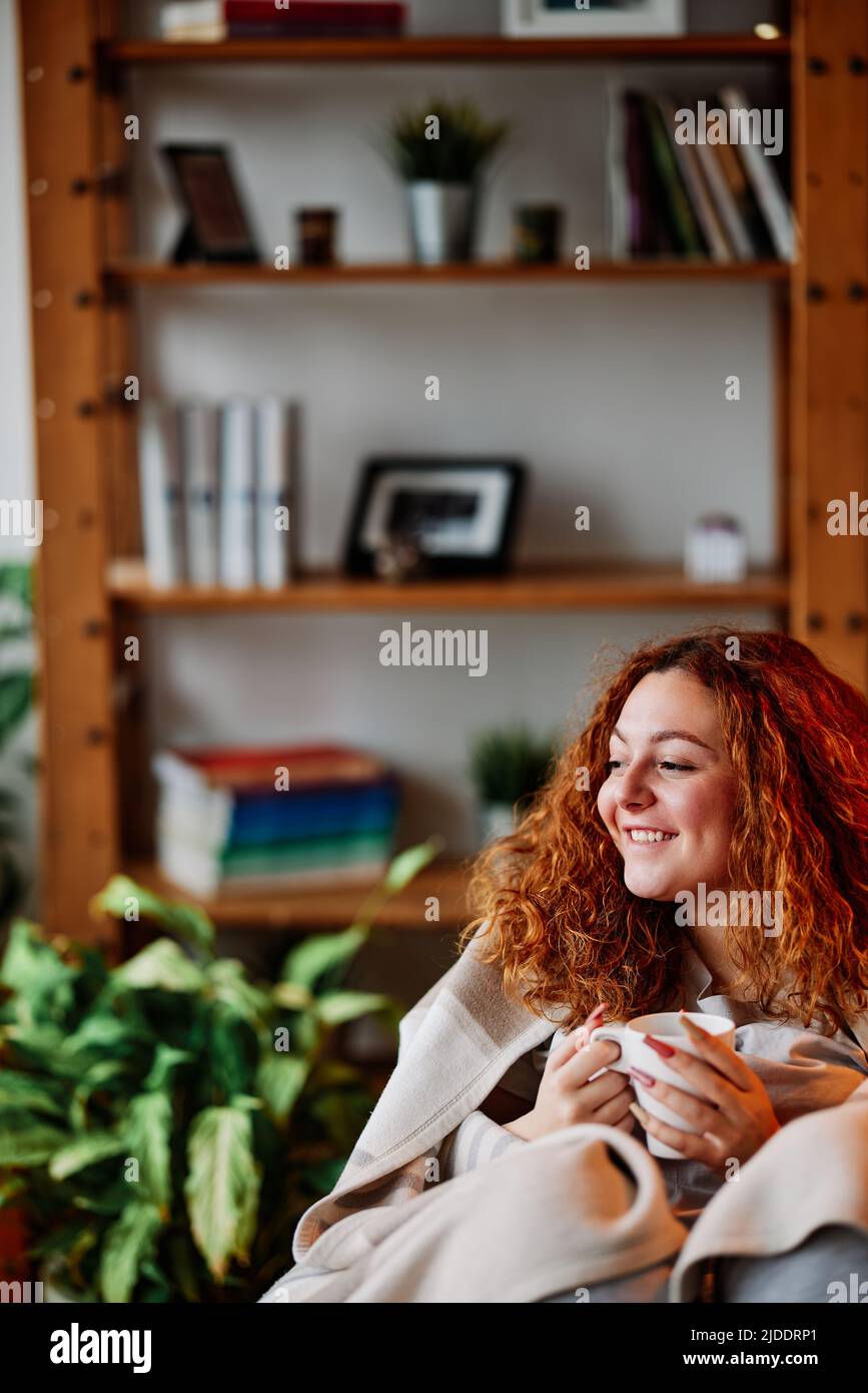Un caffè rosso che beve a casa al mattino. Una ragazza felice zenzero seduta a casa, tenendo una bella tazza di caffè caldo del mattino e godendo gratuitamente tim Foto Stock