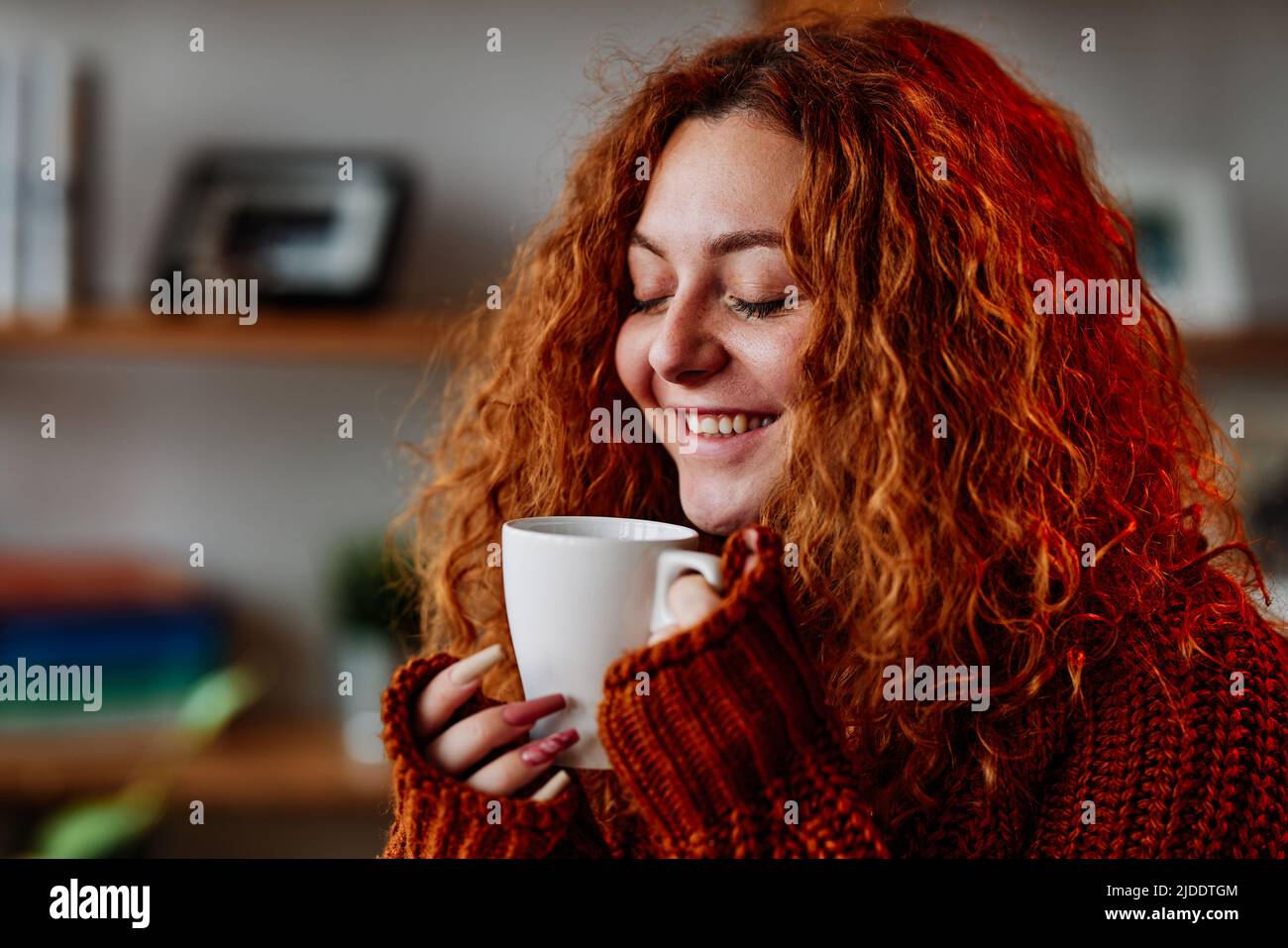 Una ragazza carina zenzero con capelli ricci è seduta nella sedia a casa al mattino e bere il suo caffè. Foto Stock