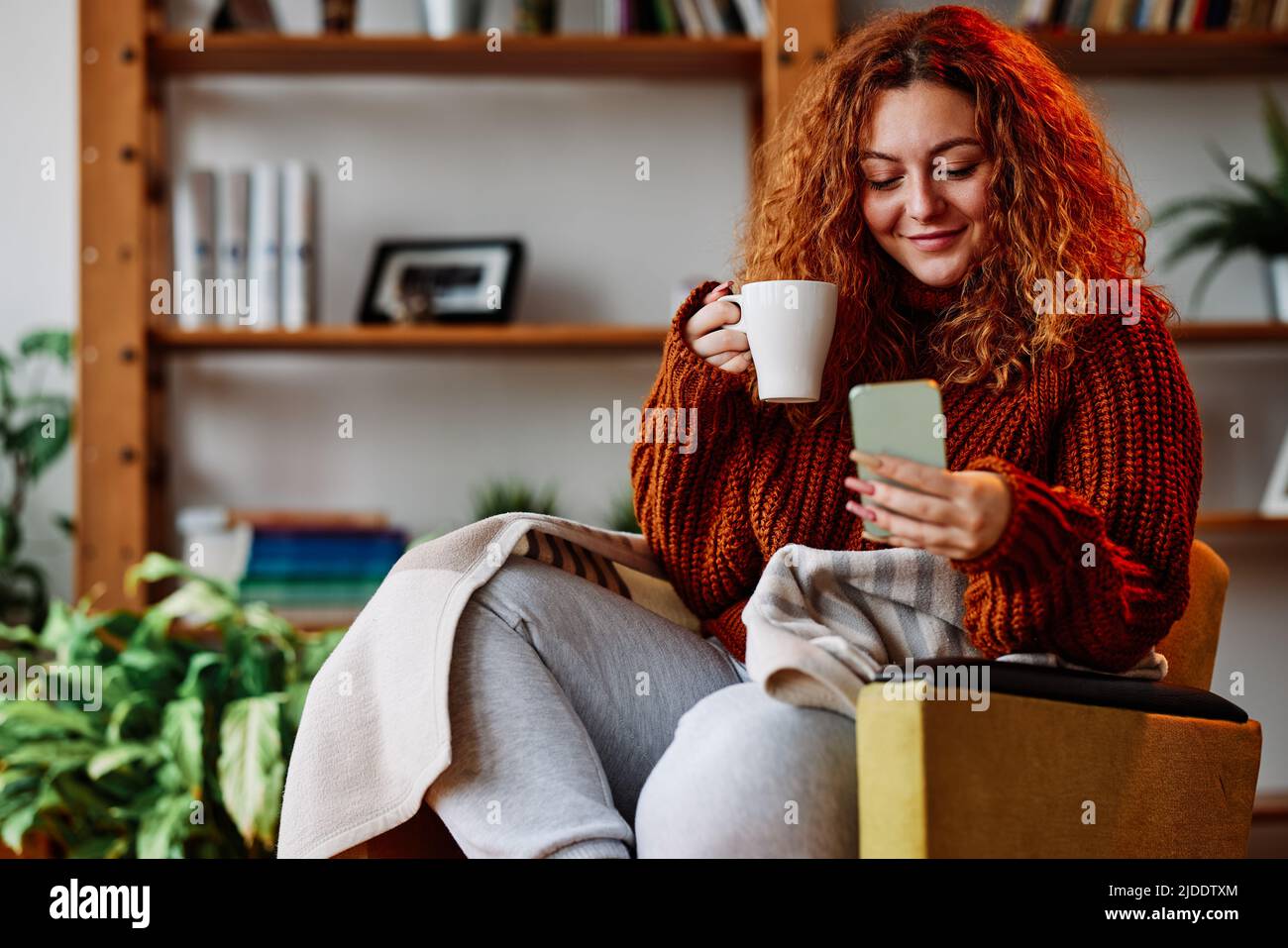 Una ragazza carina zenzero con capelli ricci è seduta nella sedia a casa al mattino, SMS messaggi sul telefono e bere il suo caffè. Foto Stock