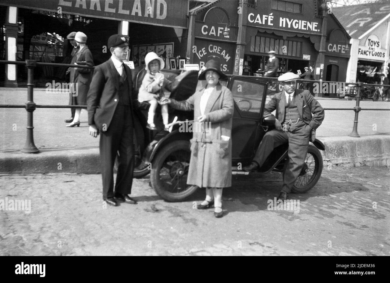 1930, storica, una famiglia che si trova accanto alla loro auto per una foto su una stradina acciottolata di pietra sul lungomare di Cleethorpes, Lincolnshire, Inghilterra, Regno Unito. Tutti i membri della famiglia sono formalmente vestiti e indossando cappelli, che per un viaggio al mare era la norma in questo periodo. Foto Stock