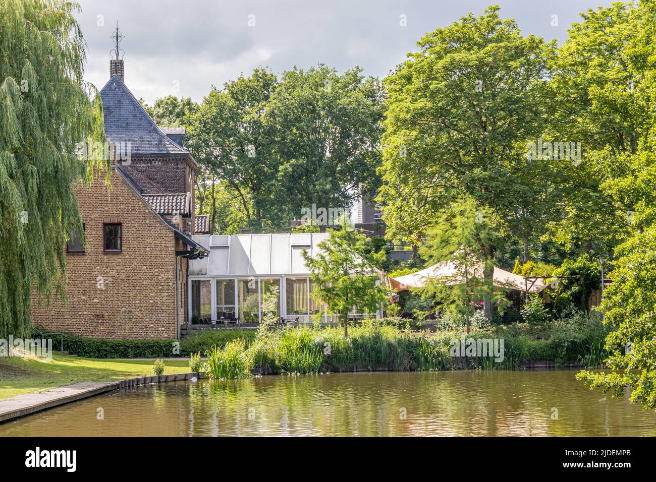 Paesaggio urbano, stagno in un parco pubblico circondato da alberi verdi lussureggianti, casa con pareti di mattoni, tetto grigio a falce con una terrazza sullo sfondo, SPR soleggiato Foto Stock