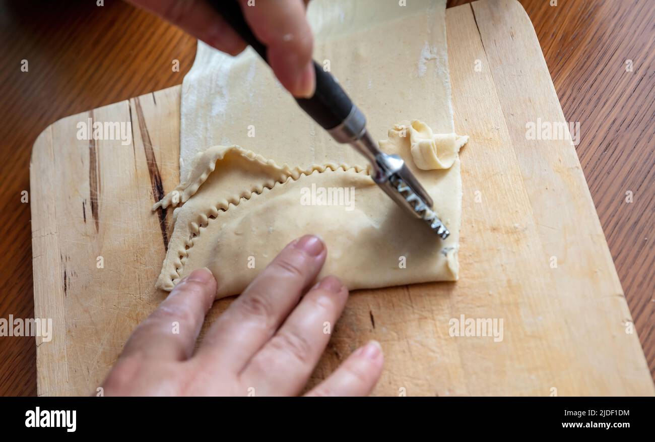 Kalitsounia preparazione tradizionale delle torte cretese. Mano femmina con una ruota di pasta frolla tagliando pasta fatta in casa farcita con formaggio di siero di latte dolce ed erbe, chiudere Foto Stock