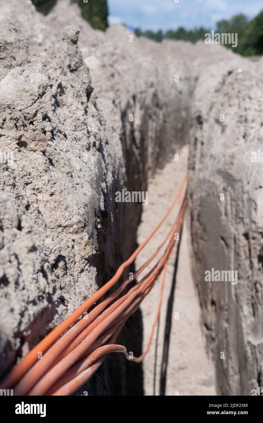 Un fascio di cavi in fibra ottica arancione si trova in una trincea scavata nel terreno in una strada. Mettere a fuoco i cavi in basso a sinistra, sfondo sfocato della Th Foto Stock