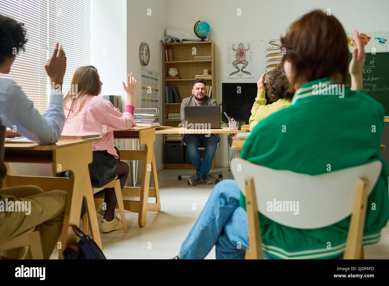 Insegnante di anatomia matura e fiducioso seduto sul posto di lavoro e porre domande agli studenti delle scuole superiori con mani rialzate a lezione Foto Stock