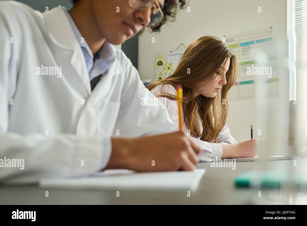 Giovane ragazza in labcoat facendo note in copybook durante il lavoro di laboratorio a lezione di chimica mentre si siede accanto al suo compagno di classe Foto Stock