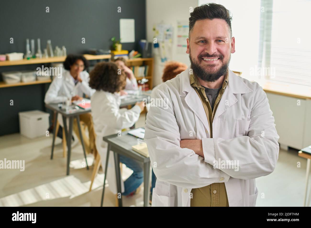 Felice bearded insegnante maturo di chimica in labcoat che attraversa le sue braccia sul petto e guardando la macchina fotografica contro il gruppo di studenti adolescenti Foto Stock