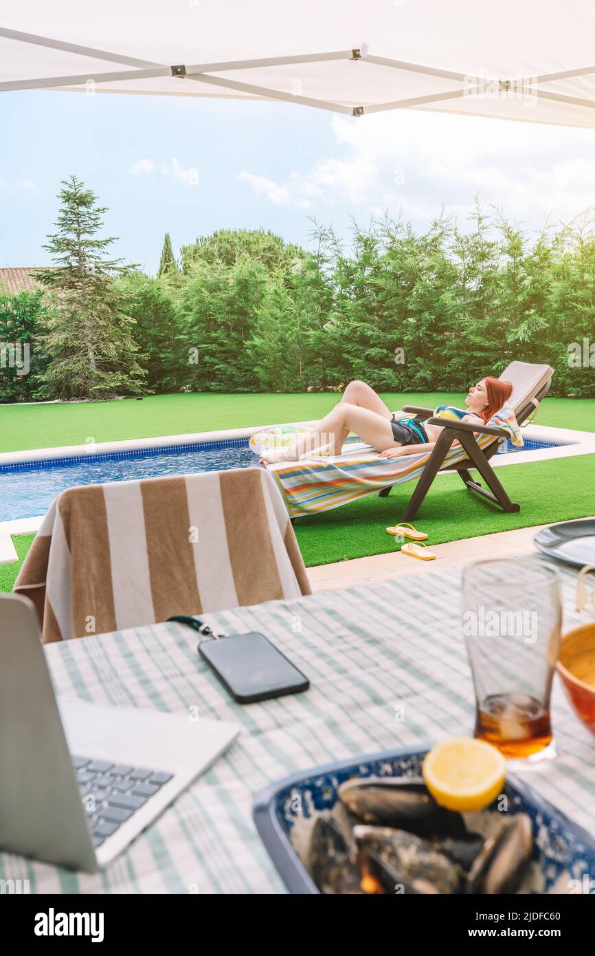 Scena di un tavolo sulla terrazza di una casa in estate con una donna sdraiata presso la piscina sullo sfondo. Scena estiva, sala da pranzo all'aperto, spazio per fotocopie e. Foto Stock
