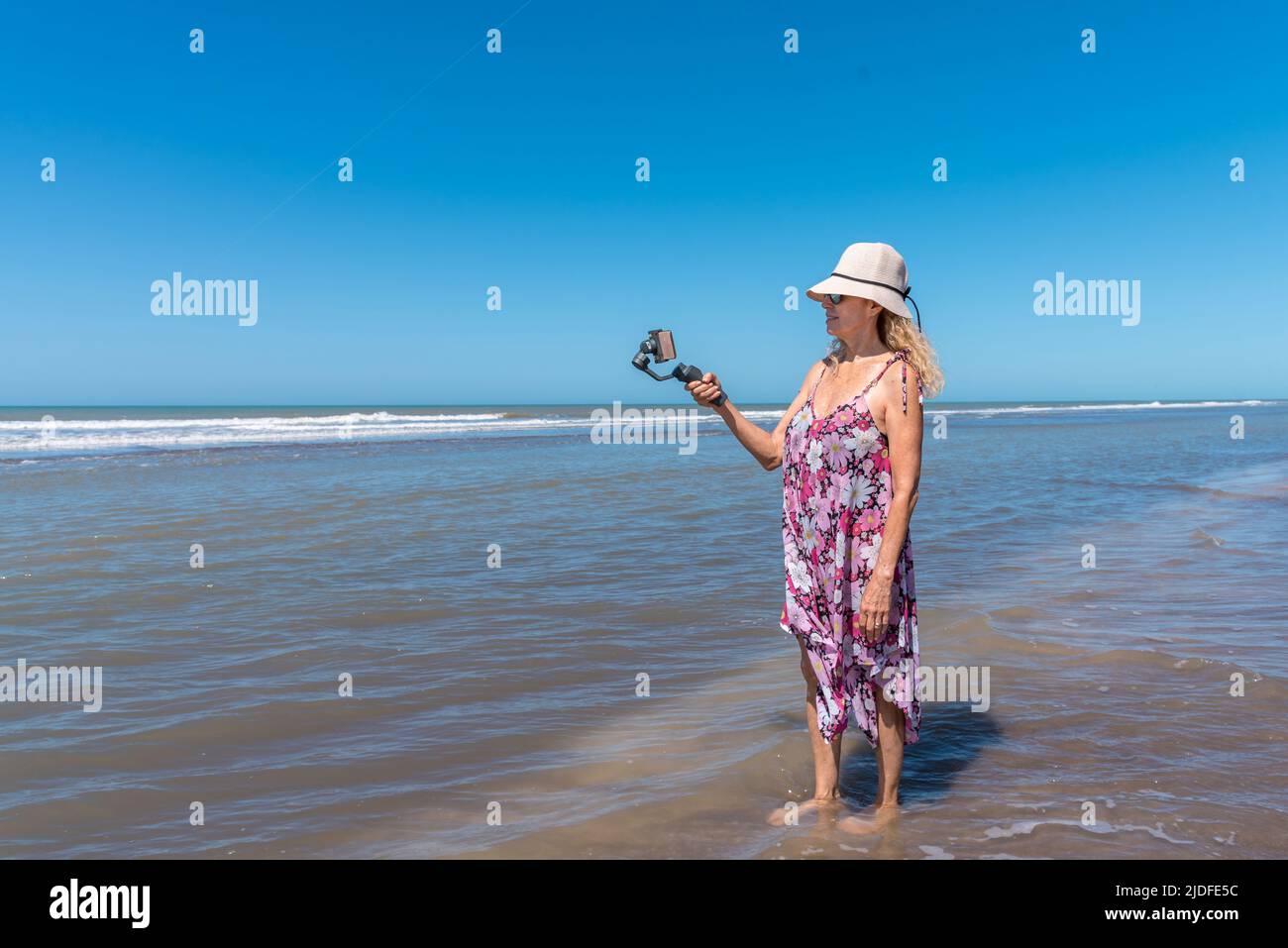 Donna bionda in estate vestire in piedi sull'acqua del mare usando un mobile attaccato su un gimbal. Foto Stock
