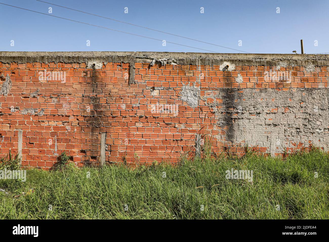 Muro muto con intonaco e alcuni mattoni esposti, con segni di tempo, evidenziandone la texture, alla base un cespuglio verde e sopra il cielo blu con wir Foto Stock