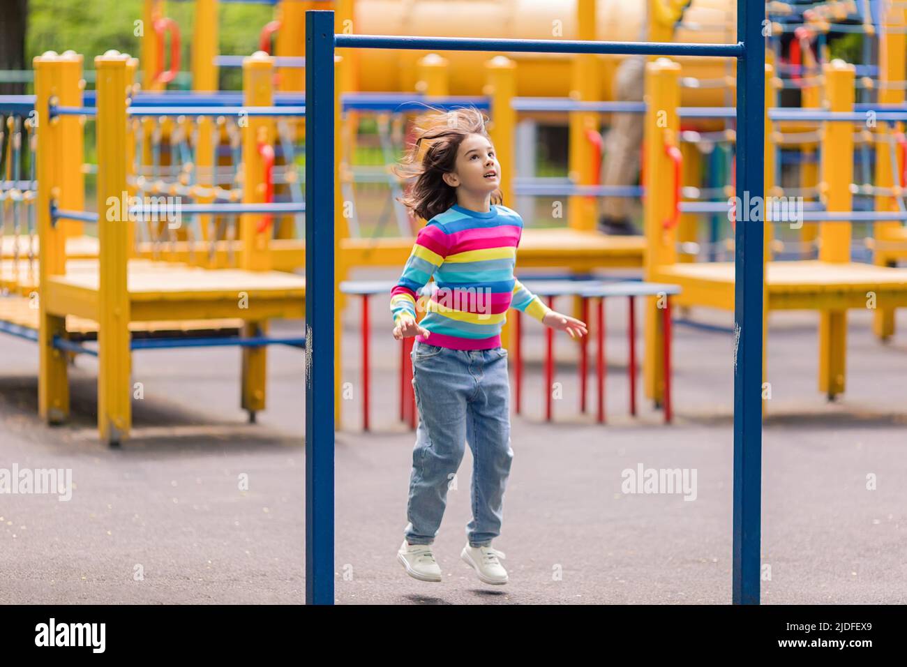 Una bambina di 5-6 anni, in un ponticello luminoso a strisce, salta sul parco giochi, nel parco Foto Stock