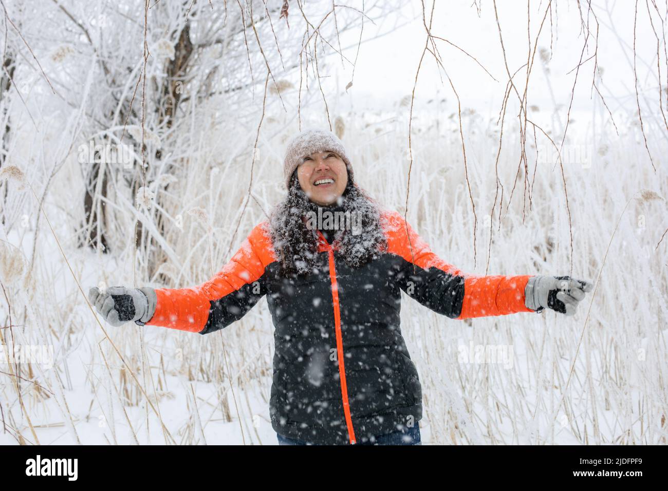 Donna beatful di mezza età con le braccia tese e faccia guardando in su sorridendo con i denti a piedi nella foresta nevosa con la neve che cade su lei. Magia Foto Stock