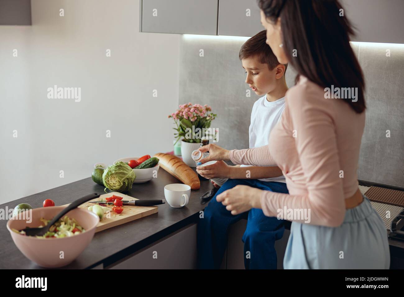 Dietro la madre caucasica che insegnava al ragazzino a cucinare. Figlio con mamma che versa il latte in una tazza. Foto Stock