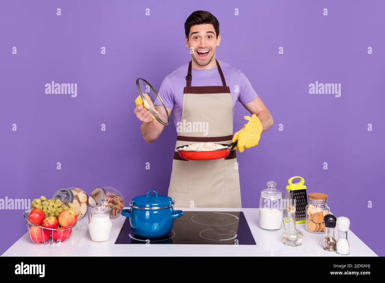 Foto di divertente ragazzo eccitato indossare grembiule grigio tenendo aperta padella cottura piatto gustoso isolato viola sfondo di colore Foto Stock