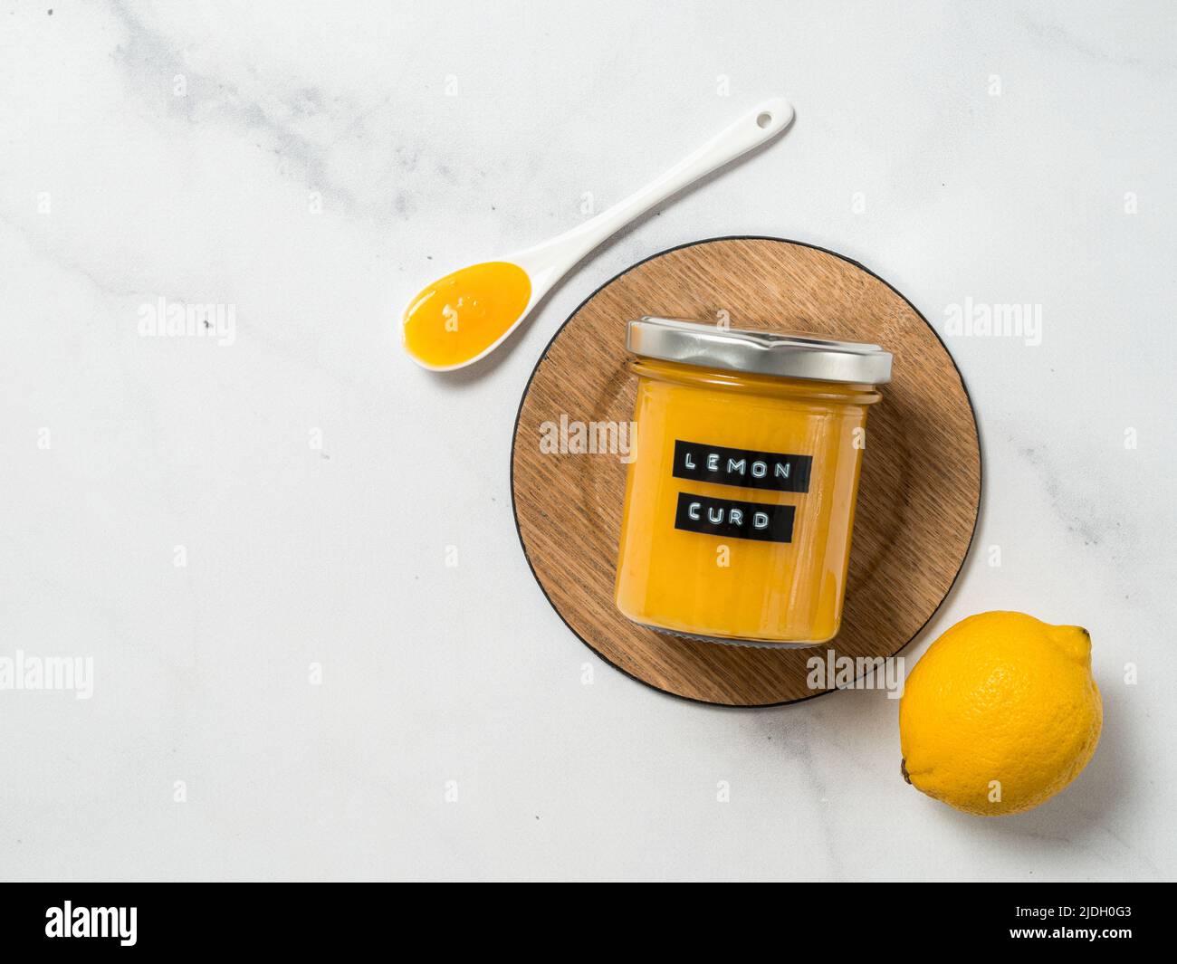 Deliziosa cagliata al limone fatta in casa. Vasetto di vetro con cagliata di limone e cucchiaio pieno su sfondo di marmo bianco. Etichetta adesiva nera con il testo Lemon Curd sul vaso. Vista dall'alto o disposizione piatta. Spazio di copia Foto Stock