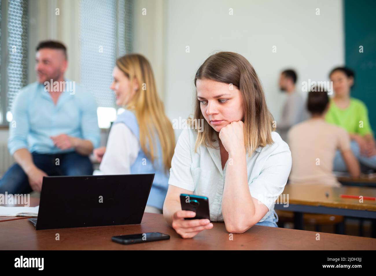 Ragazza turbata seduta a tavola in auditorium durante la pausa sullo sfondo con gli studenti Foto Stock