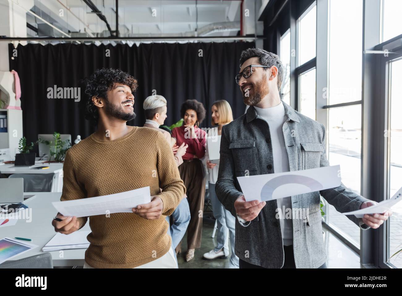 ridere gli uomini interrazziali con carte che si guardano l'un l'altro in agenzia pubblicitaria Foto Stock