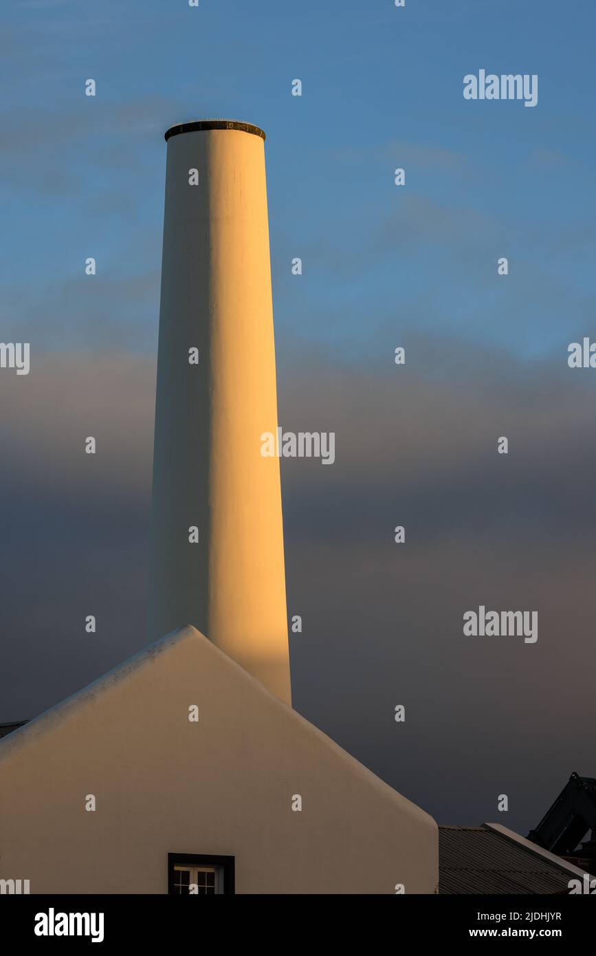 la luce del sole al tramonto illumina un lato di un alto camino bianco di fabbrica sullo sfondo cielo blu chiaro sopra le nuvole scure Foto Stock