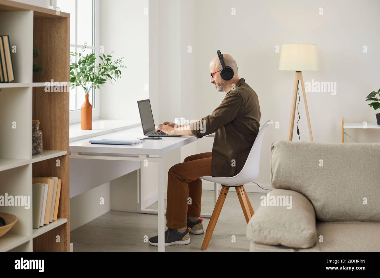 Uomo adulto sorridente che ascolta la musica con le cuffie mentre lavora sul laptop a casa. Foto Stock