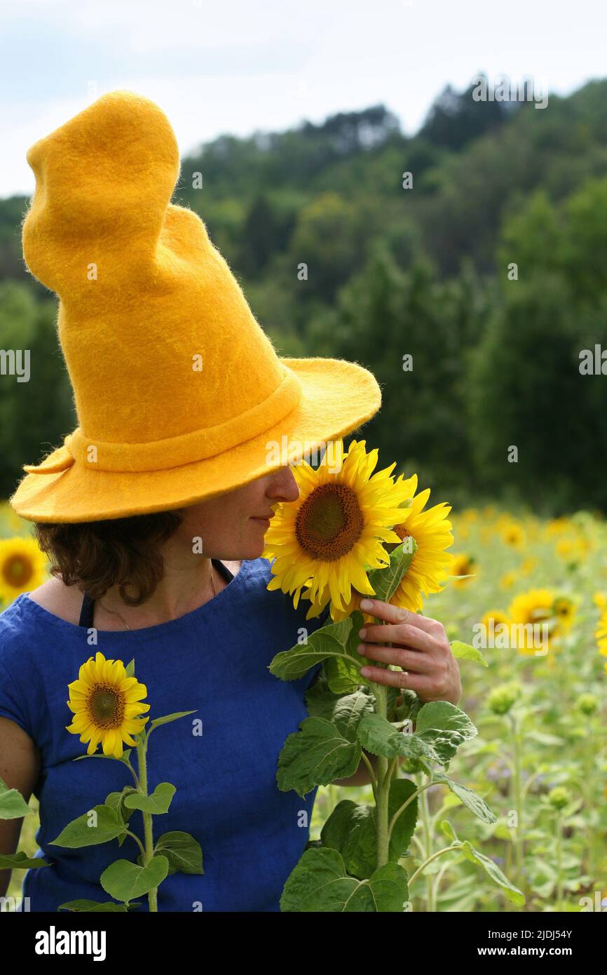 Donna con capelli ricci corti marroni vestito blu reale e cappello giallo grande corona alta con girasole odore di brim largo. Passeggiate nel campo di girasole Foto Stock