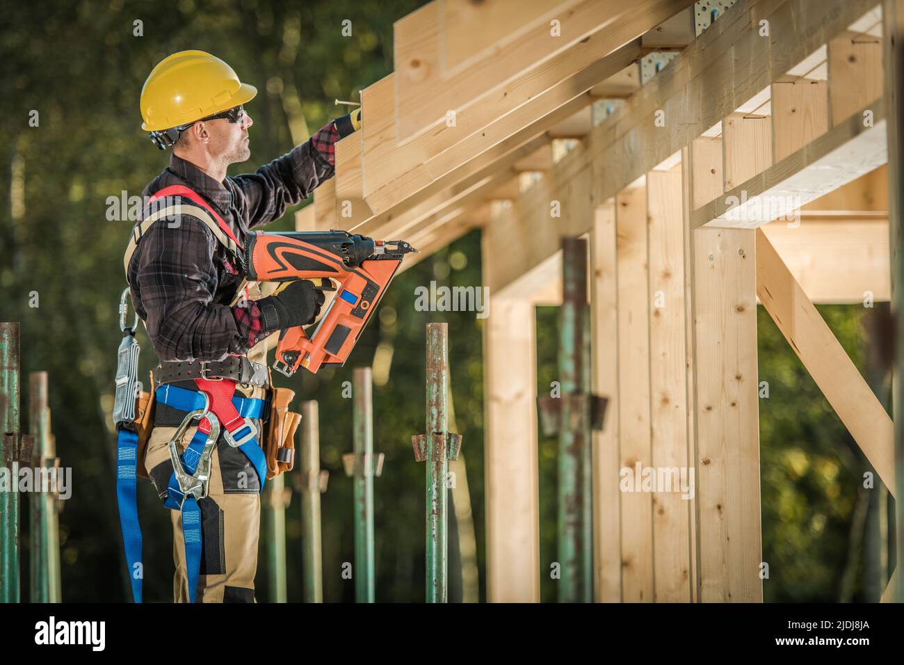 Carpenter caucasico professionalmente equipaggiato al suo 40s lavorando sulla finitura del tetto in legno skeleton utilizzando Nail Gun Construction Tool. Costruzione si Foto Stock
