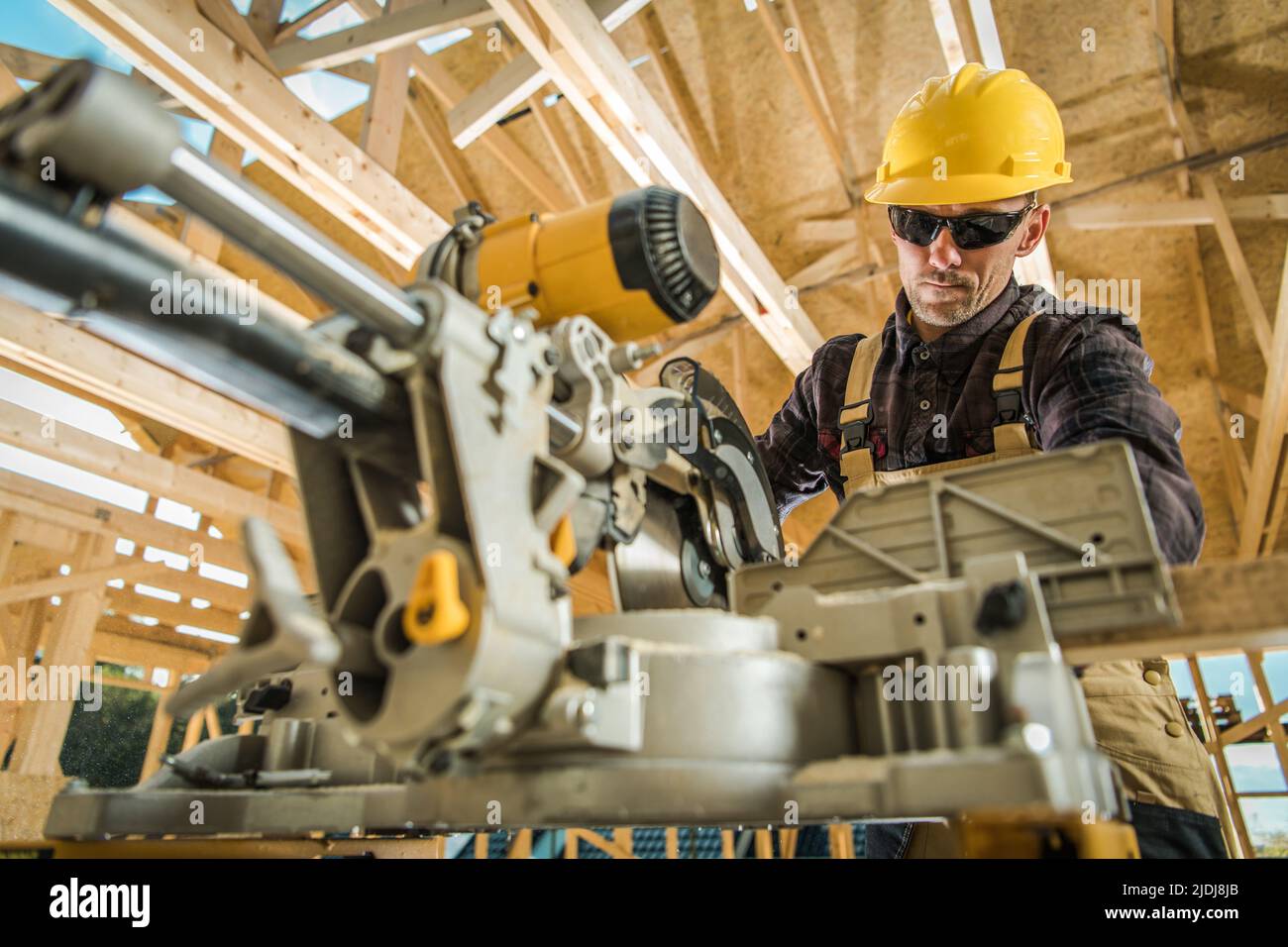 Lavoratore caucasico che utilizza una sega da tavolo mentre lavora alla costruzione dello scheletro di legno di un edificio residenziale. Cantiere sfondo. L Foto Stock