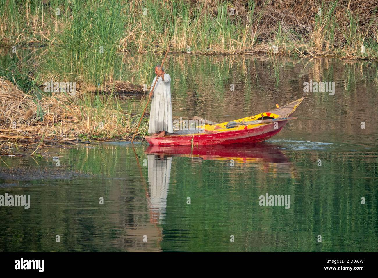Un pescatore solista del Nilo che tiene il bastone lungo in piedi in barca di pesca di legno dipinta luminosamente in una piccola insenatura con acqua e riflessi delicatamente increspati Foto Stock