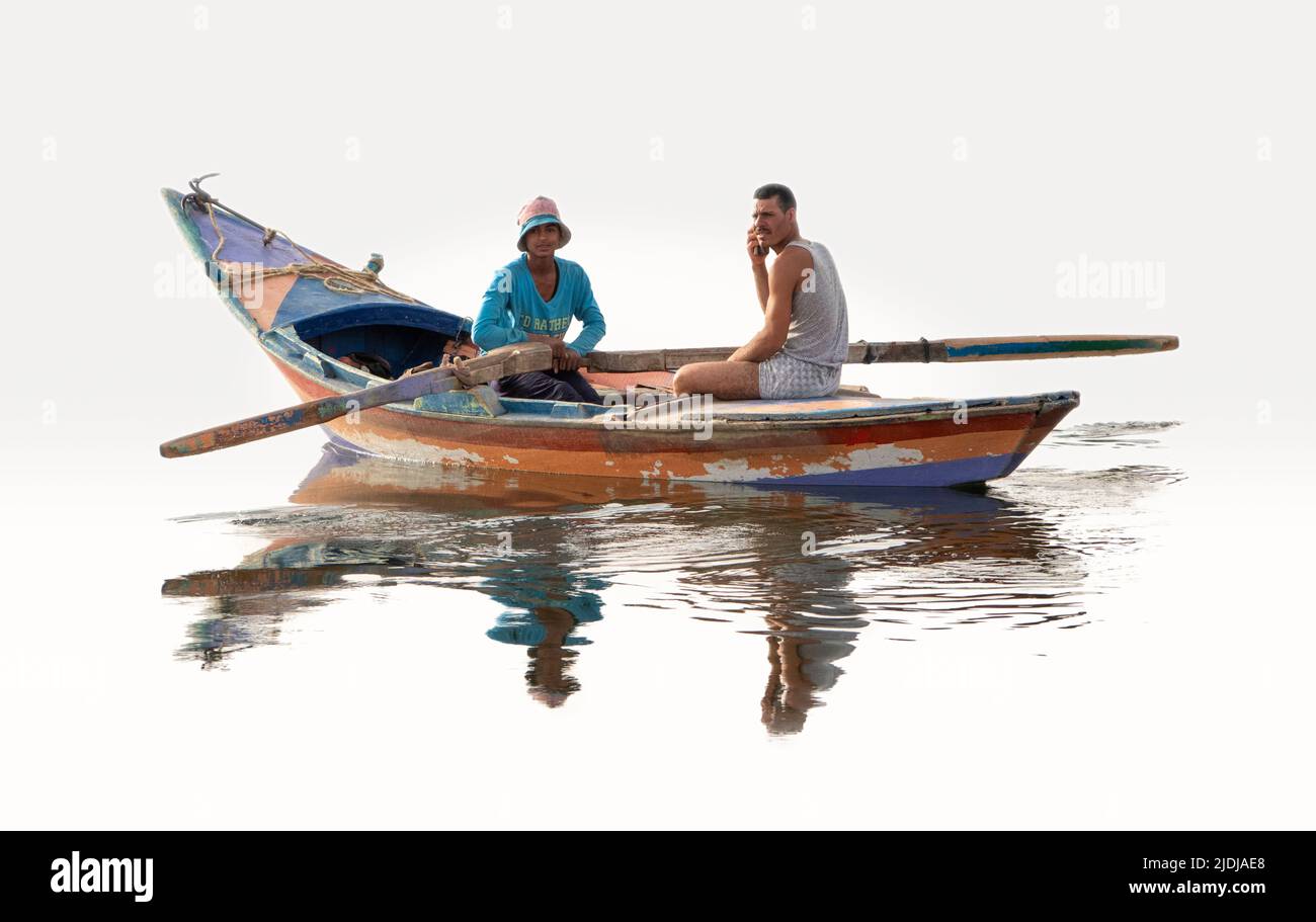 Due giovani uomini, uno canottaggio e uno sul telefono cellulare in una barca di legno dipinta in modo brillante, perfettamente riflessa in specchi d'acqua di superficie Foto Stock