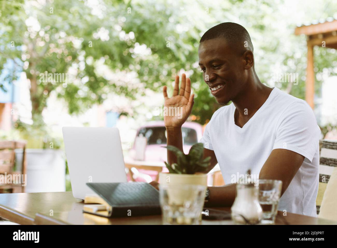 Uomo multirazziale sorridente e felice con un computer portatile, sollevando il braccio, salutando qualcuno, guardando il monitor. Incontro del webinar sull'e-learning Foto Stock