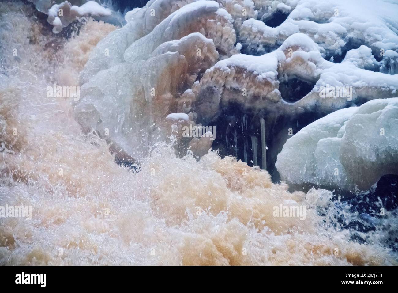 Cataratta a cascata in montagne forestali. La sorgente e la massa di acqua fangosa scongelata cade dalla scogliera tra la foresta di abete rosso innevata. Copertura delle sponde delle cascate Foto Stock