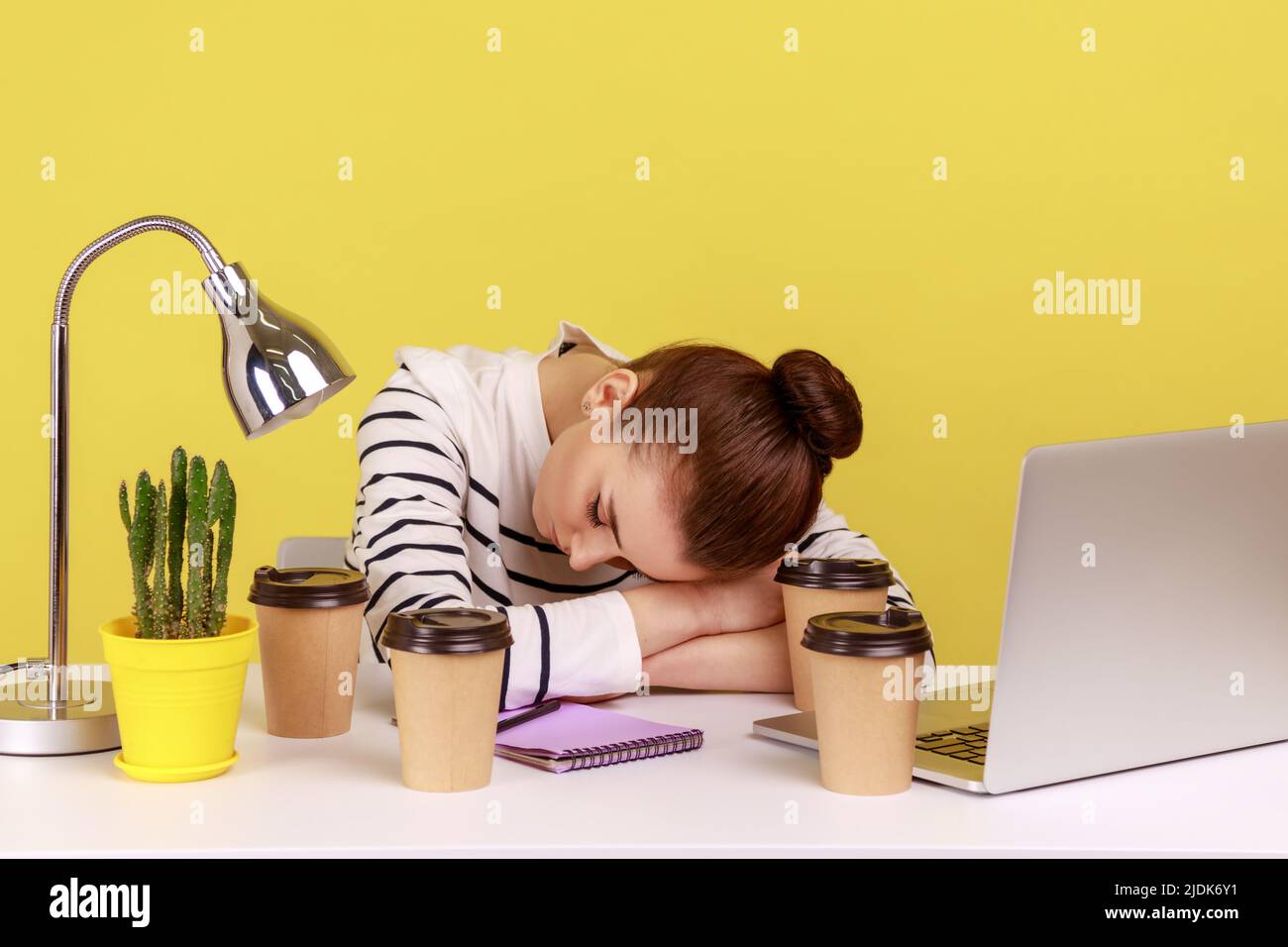 Una donna affaticata che dorme sul posto di lavoro, distesa circondata da tazze di caffè, riposa dopo il lavoro straordinario. Studio interno girato isolato su sfondo giallo. Foto Stock