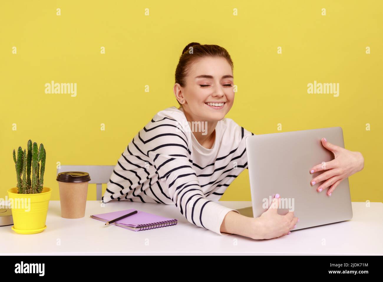 Felice donna soddisfatta che abbraccia il laptop con il sorriso che ha piacevole colloquio sulla videochiamata, intervista di lavoro in linea, conferenza dall'ufficio domestico. Studio interno girato isolato su sfondo giallo. Foto Stock