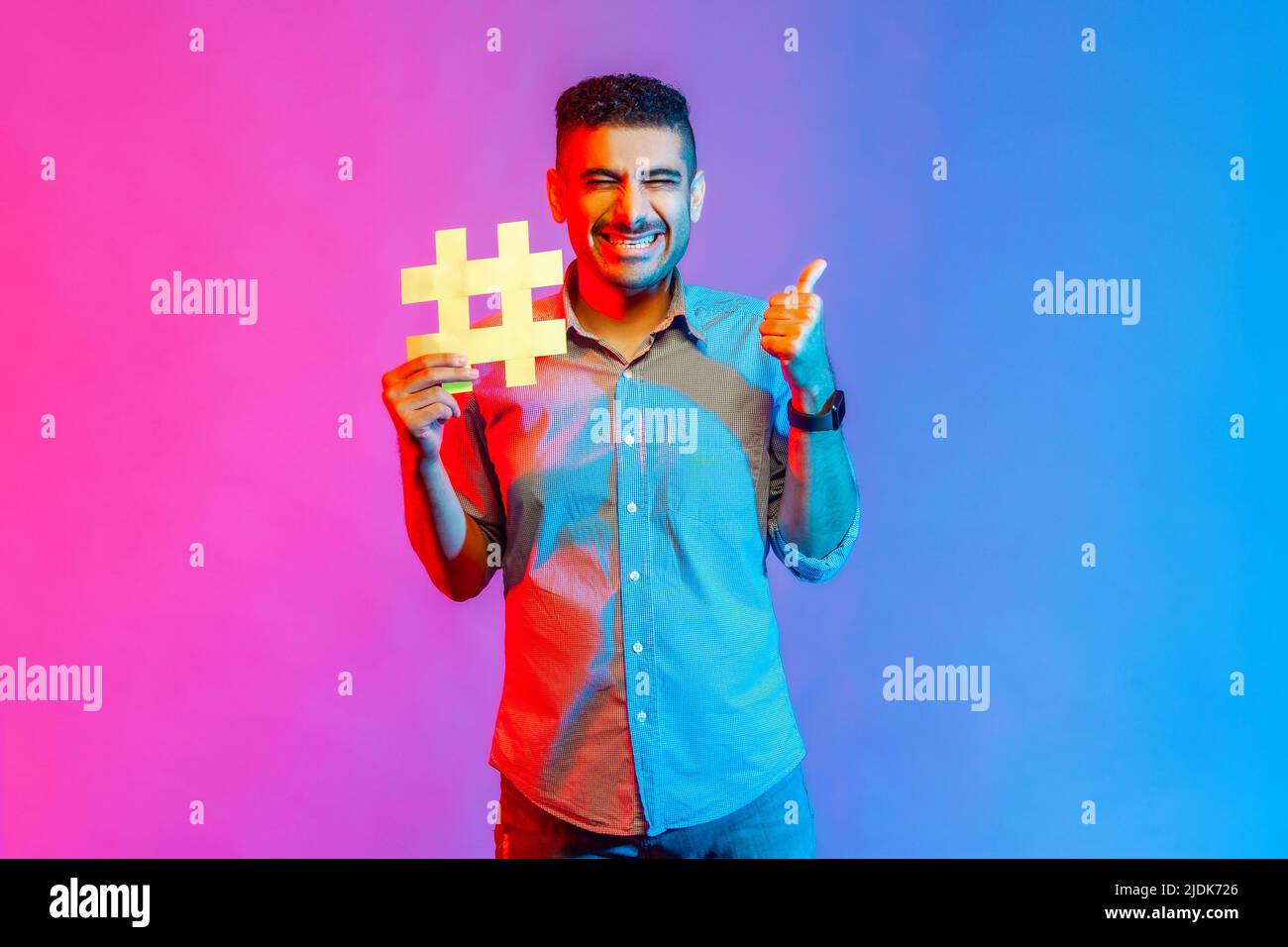 Ritratto di uomo in camicia con sorriso toothy, tenendo hashtag simbolo, mostrando pollice in su, consigliando i soggetti popolari, tendenze del Internet. Studio interno girato isolato su colorato sfondo di luce al neon. Foto Stock