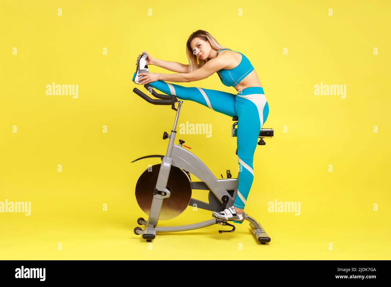 Ritratto di giovane adulto bella donna sportiva stretching gamba prima di allenarsi su simulatore di bicicletta, guardando via, indossando abbigliamento sportivo blu. Studio interno girato isolato su sfondo giallo. Foto Stock