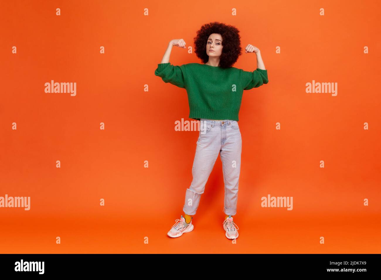 Ritratto a tutta lunghezza di donna con acconciatura Afro in braccia di sollevamento maglione verde che mostra bicipite come metafora di potere femminile e indipendenza, femminismo. Studio interno girato isolato su sfondo arancione. Foto Stock