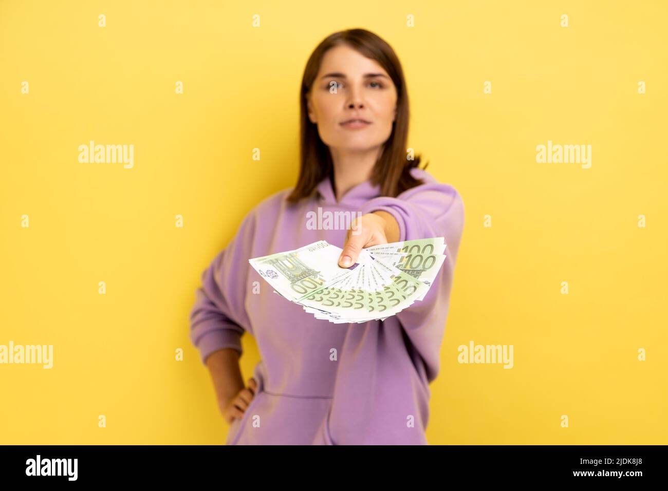 Ritratto di giovane donna fiduciosa ricca con capelli scuri che tengono fuori le banconote in euro alla macchina fotografica, offrendo soldi, indossando la felpa con cappuccio viola. Studio interno girato isolato su sfondo giallo. Foto Stock