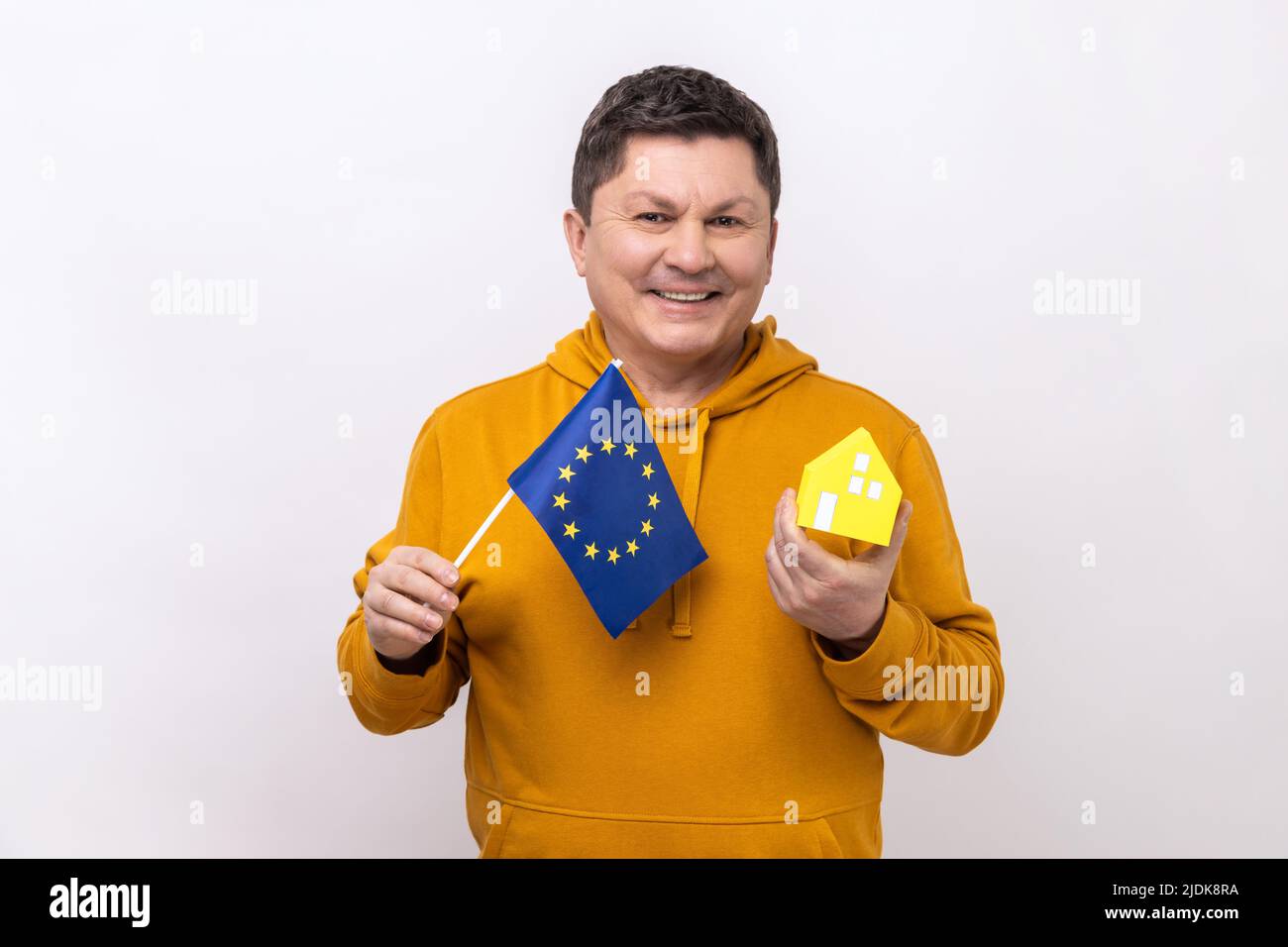 Uomo positivo di mezza età che detiene bandiera europea e casa di carta, sognando di comprare la sua casa in Europa, guardando la macchina fotografica, indossando una felpa con cappuccio in stile urbano. Studio interno girato isolato su sfondo bianco. Foto Stock