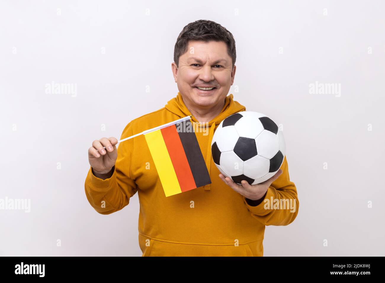 Uomo di mezza età con il sorriso toothy che tiene la bandiera della germania e calcio palla classica in bianco e nero e guardare la partita, indossando felpa con cappuccio urban style. Studio interno girato isolato su sfondo bianco. Foto Stock