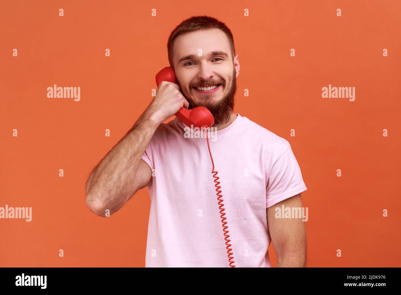 Ritratto di uomo bearded che parla il telefono fisso che tiene in mano il ricevitore, guardando la macchina fotografica con il sorriso toothy, che indossa la T-shirt rosa. Studio interno girato isolato su sfondo arancione. Foto Stock