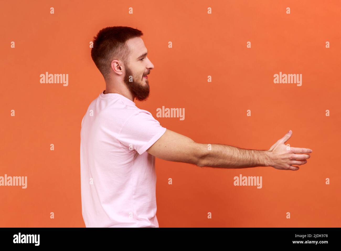 Ritratto della vista laterale di un bell'uomo portamonete positivo e amichevole che teneva la mano a fianco, dando una stretta di mano, indossando una T-shirt rosa. Studio interno girato isolato su sfondo arancione. Foto Stock