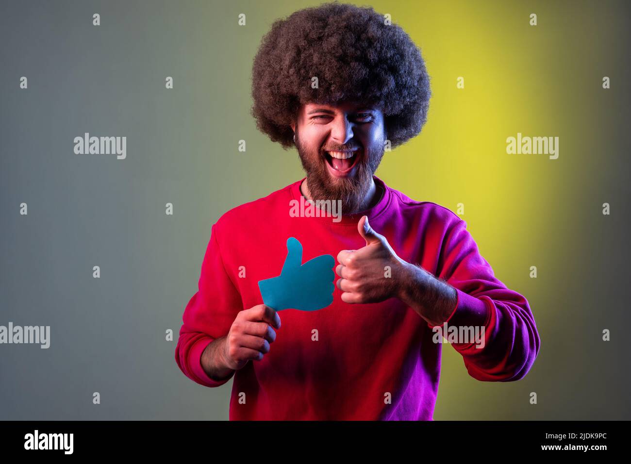 Ritratto di hipster felice uomo con acconciatura Afro che tiene come carta forma segno e mostrando pollice in su con il dito, indossando la felpa rossa. Studio interno girato isolato su colorato sfondo di luce al neon. Foto Stock