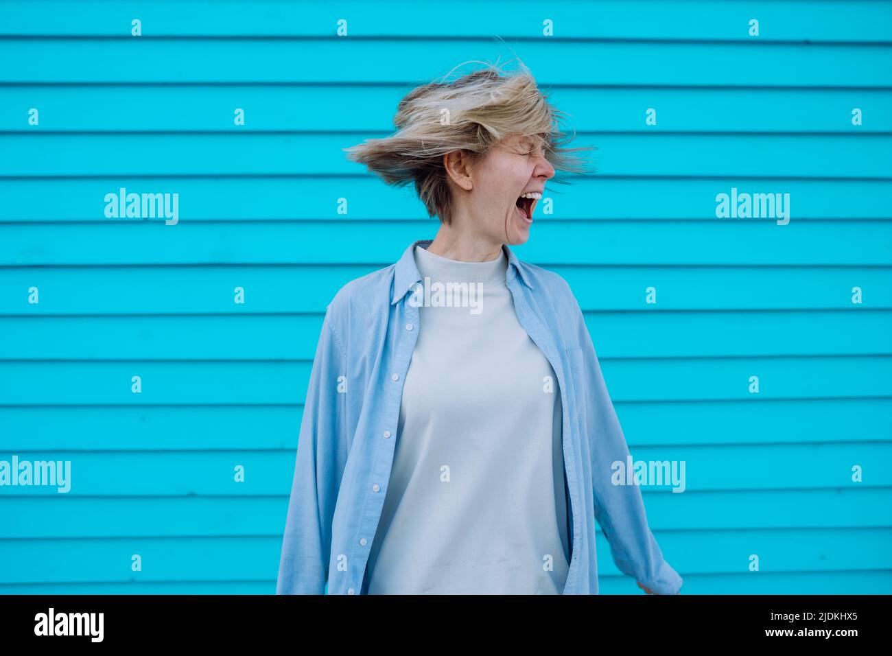 Donna pazza di mezza età con capelli volanti in piedi alla parete blu, urlando gridare, sensazione di stress, disperazione, panico. Foto Stock