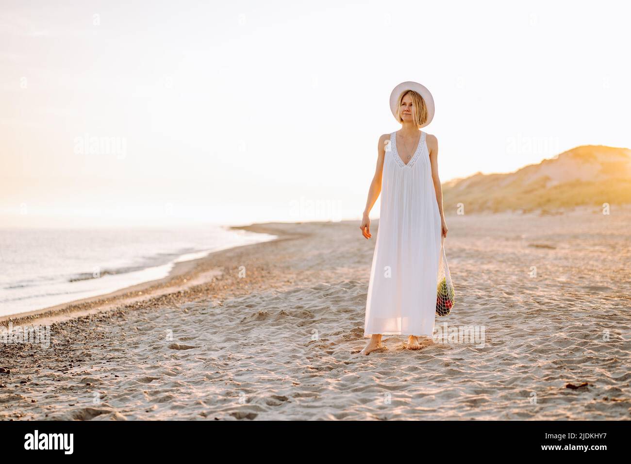 Ritratto di donna di mezza età in piedi sulla spiaggia sabbiosa vicino al mare al tramonto, portando borsa stringa piena di prodotti. Foto Stock