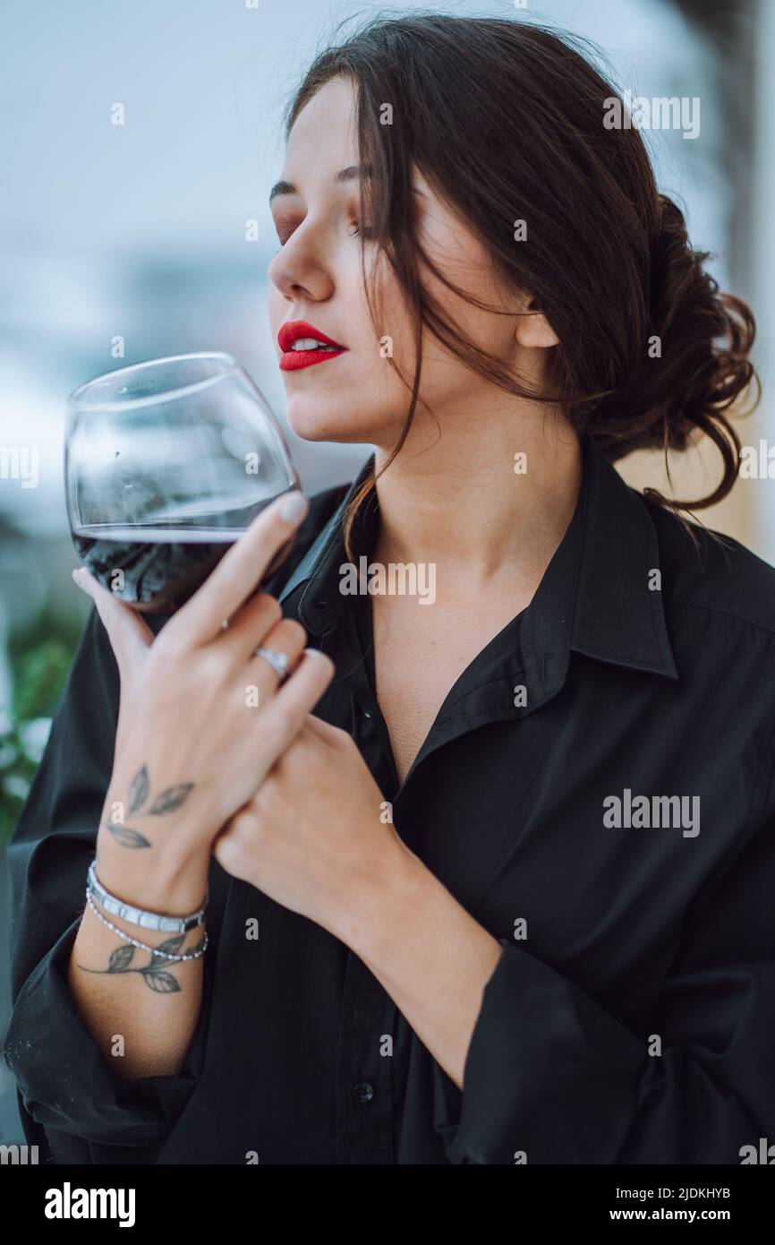 Ritratto di giovane donna splendida con capelli scuri, make-up indossando camicia nera, tenendo vetro con vino rosso, occhi chiusi. Foto Stock