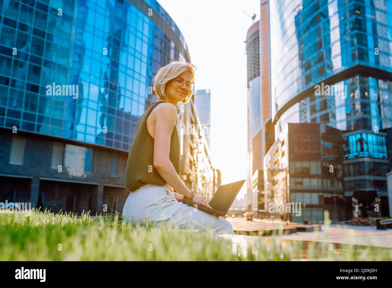 Ritratto di donna di mezza età con top verde, pantaloni bianchi, seduto nel centro della città vicino ai grattacieli che reggono un computer portatile. Foto Stock