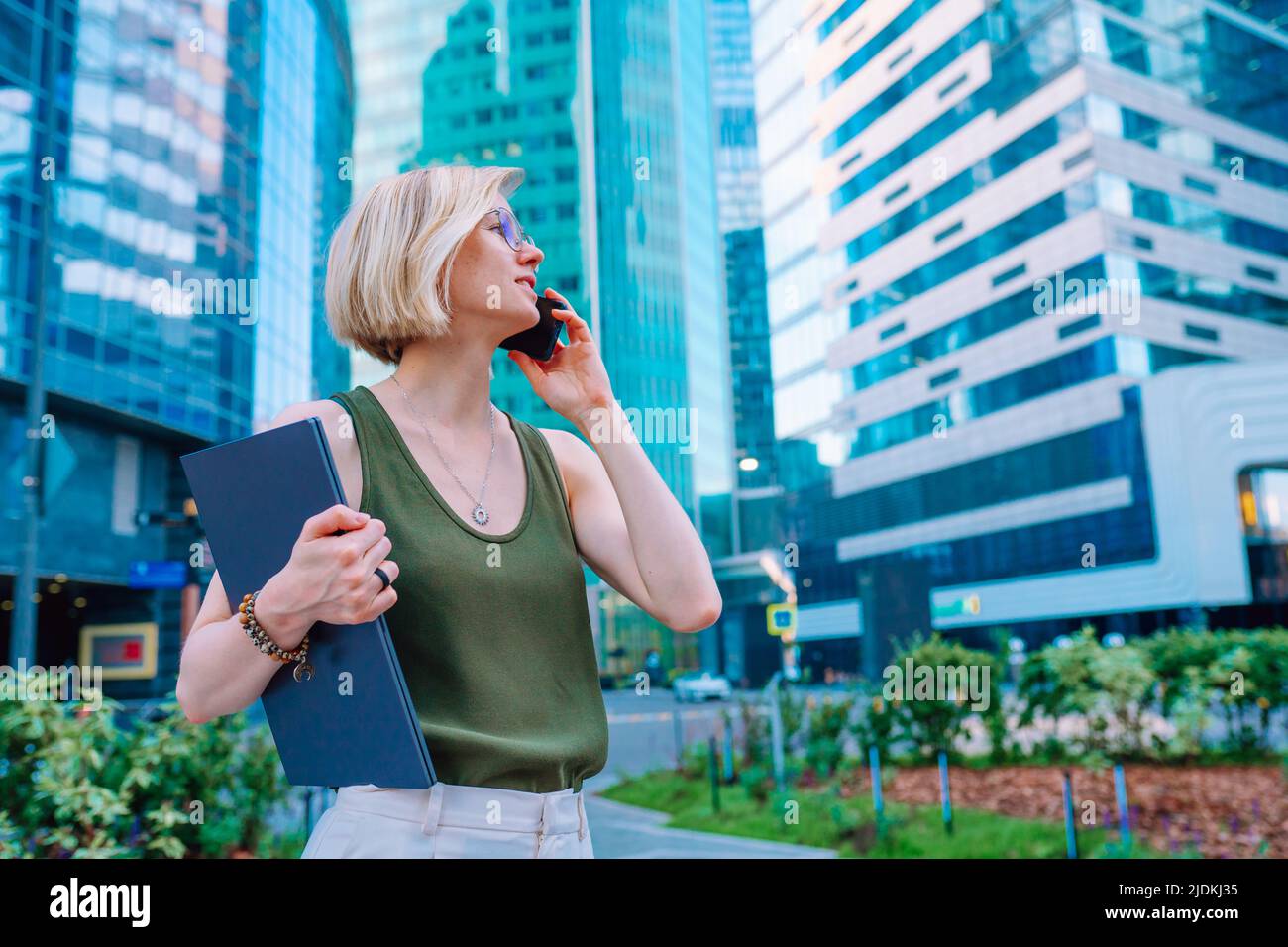 Vista laterale della donna di mezza età donna d'affari in piedi in strada nel centro della città, tenendo un laptop, parlando su smartphone. Foto Stock