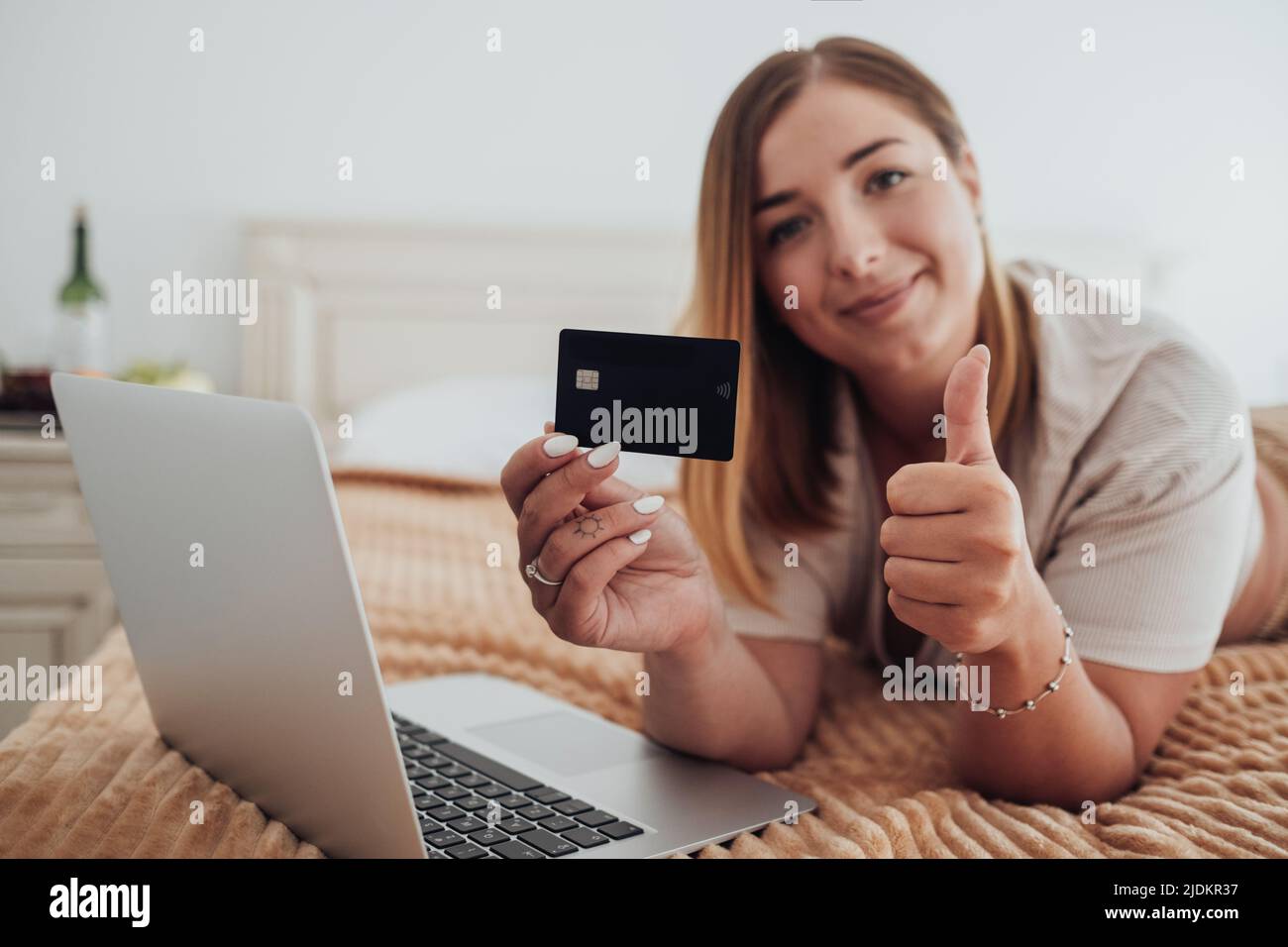 Giovane donna caucasica che tiene la carta di credito in mano e mostra il pollice in su mentre si posa sul letto della stanza dell'hotel e facendo lo shopping in linea con il laptop Foto Stock